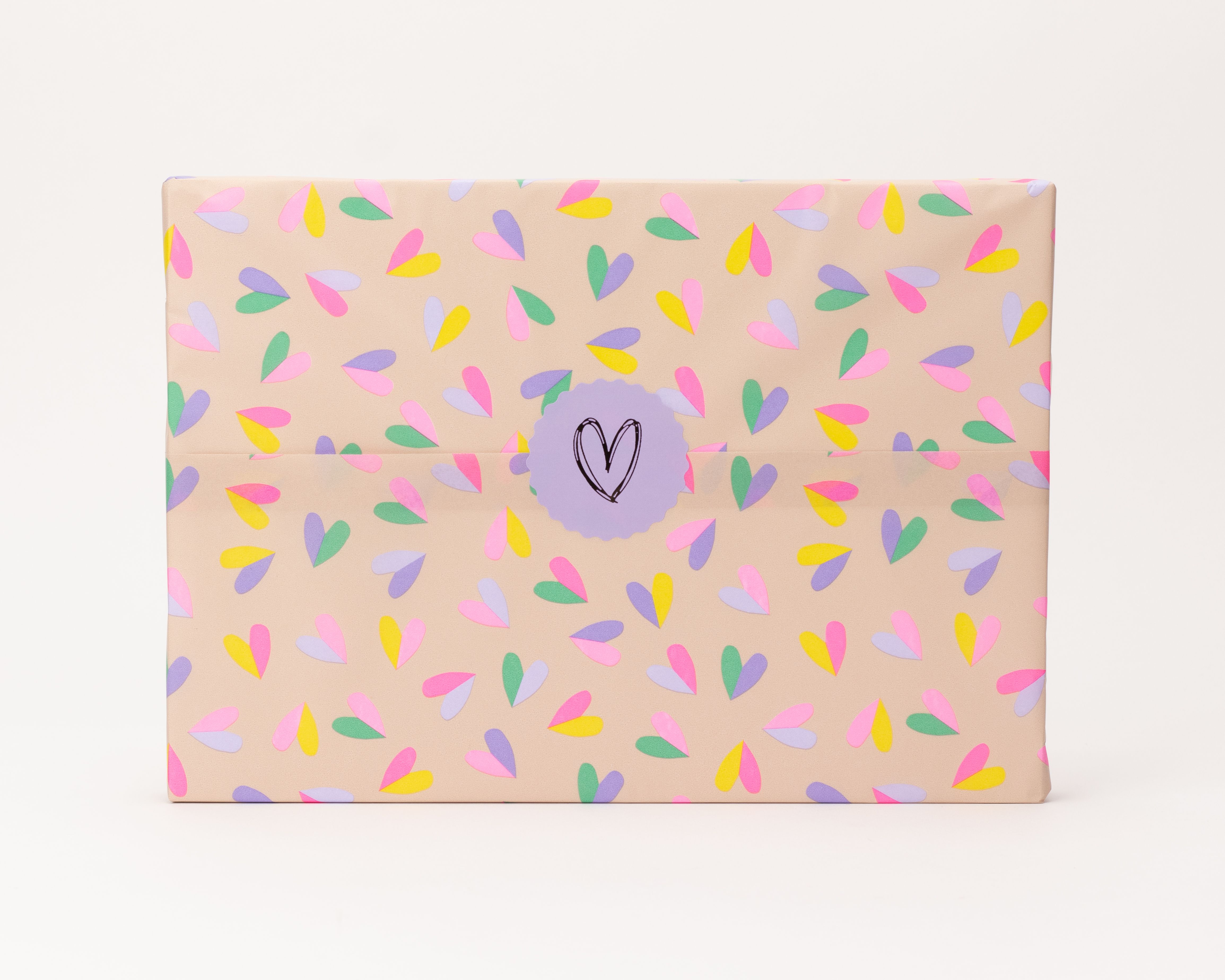 Seidenpapier in Beige mit Herzmuster in den Pastellfarben Rosa, Gelb, Lila und Mintgrün. Darauf ein runder, lila Aufkleber mit einem schwarzen, handgezeichneten Herz.