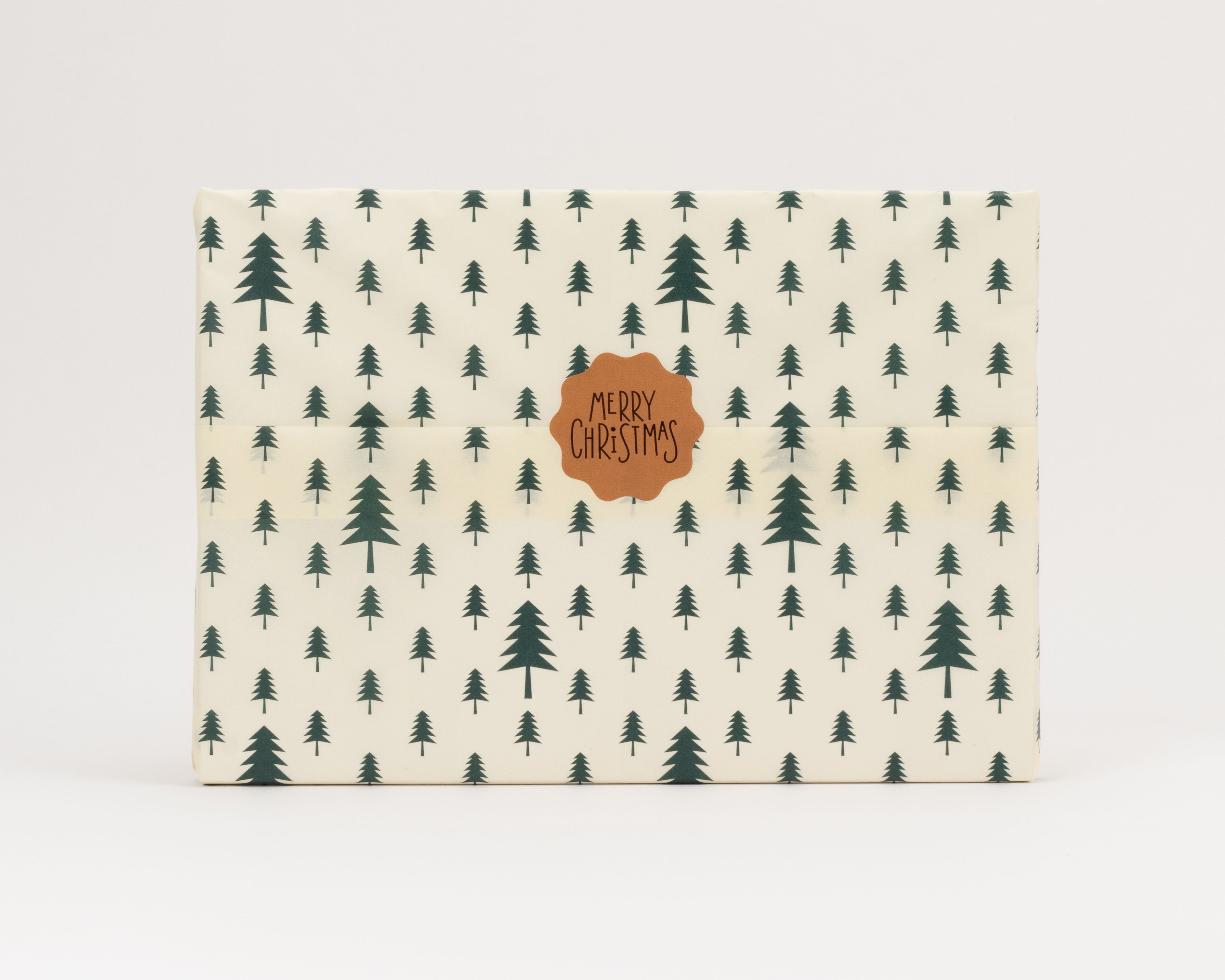 Seidenpapier mit dunkelgrünen Tannenbäumen auf cremefarbenem Grund. Darauf ein brauner Aufkleber mit der Aufschrift Merry Christmas