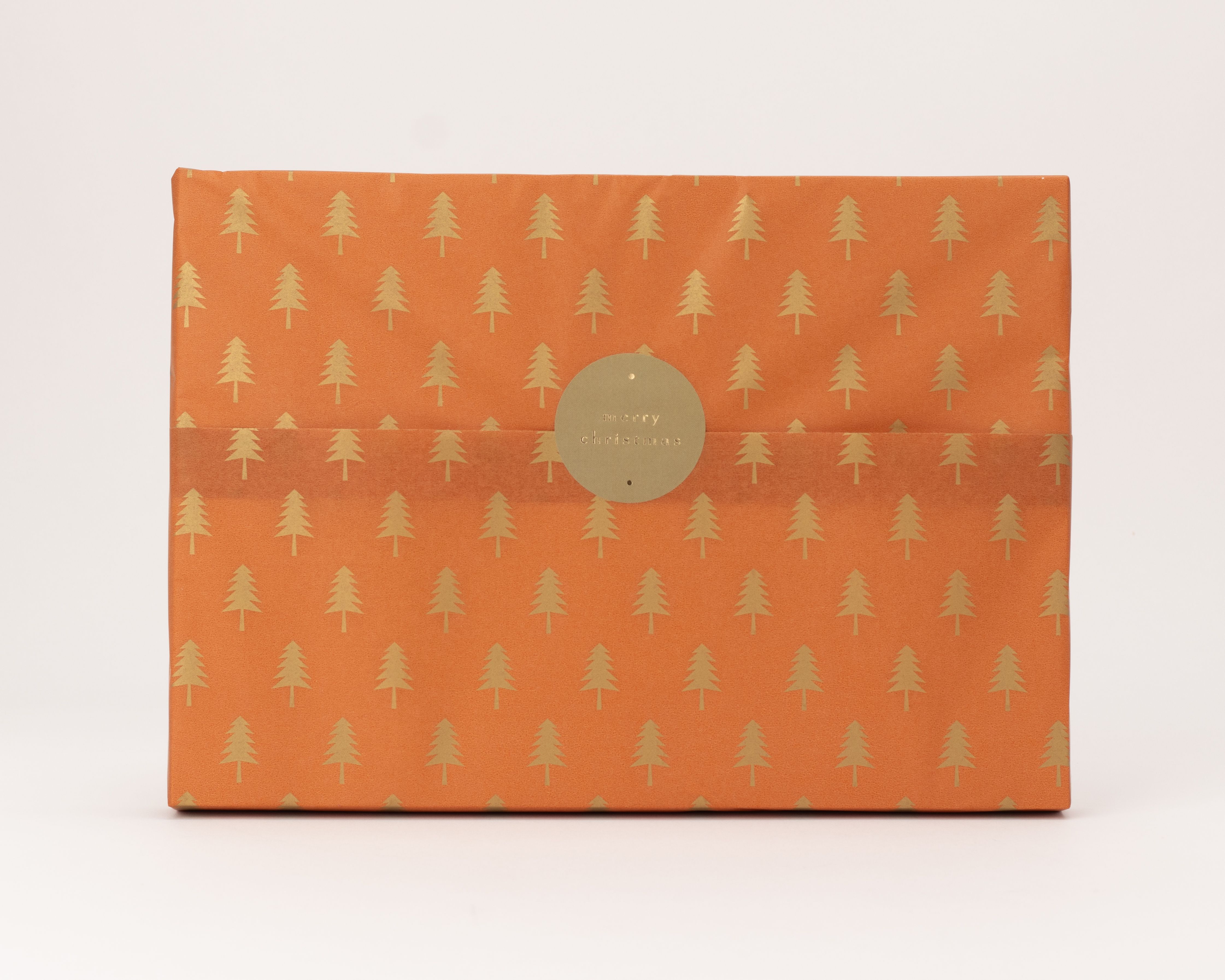 Seidenpapier in Rostfarbe mit goldenen Tannenbäumen als Muster. Darauf ein runder goldener Aufkleber mit dem Text merry christmas