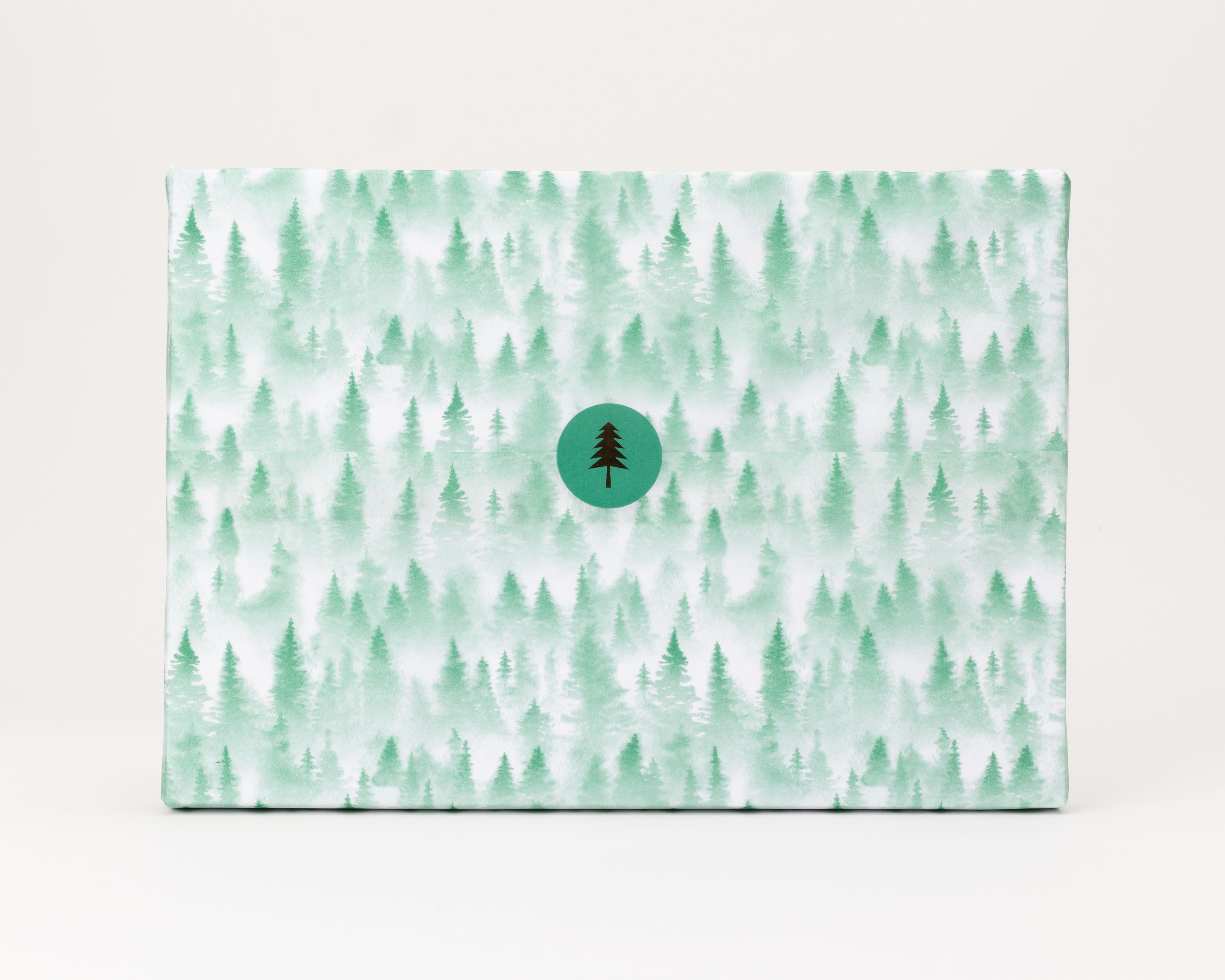 Seidenpapier mit hellgruenem Tannenwald-Muster in verschiedenen Gruentoenen. Darauf ein runder, dunkelgruener Aufkleber mit brauner Tanne