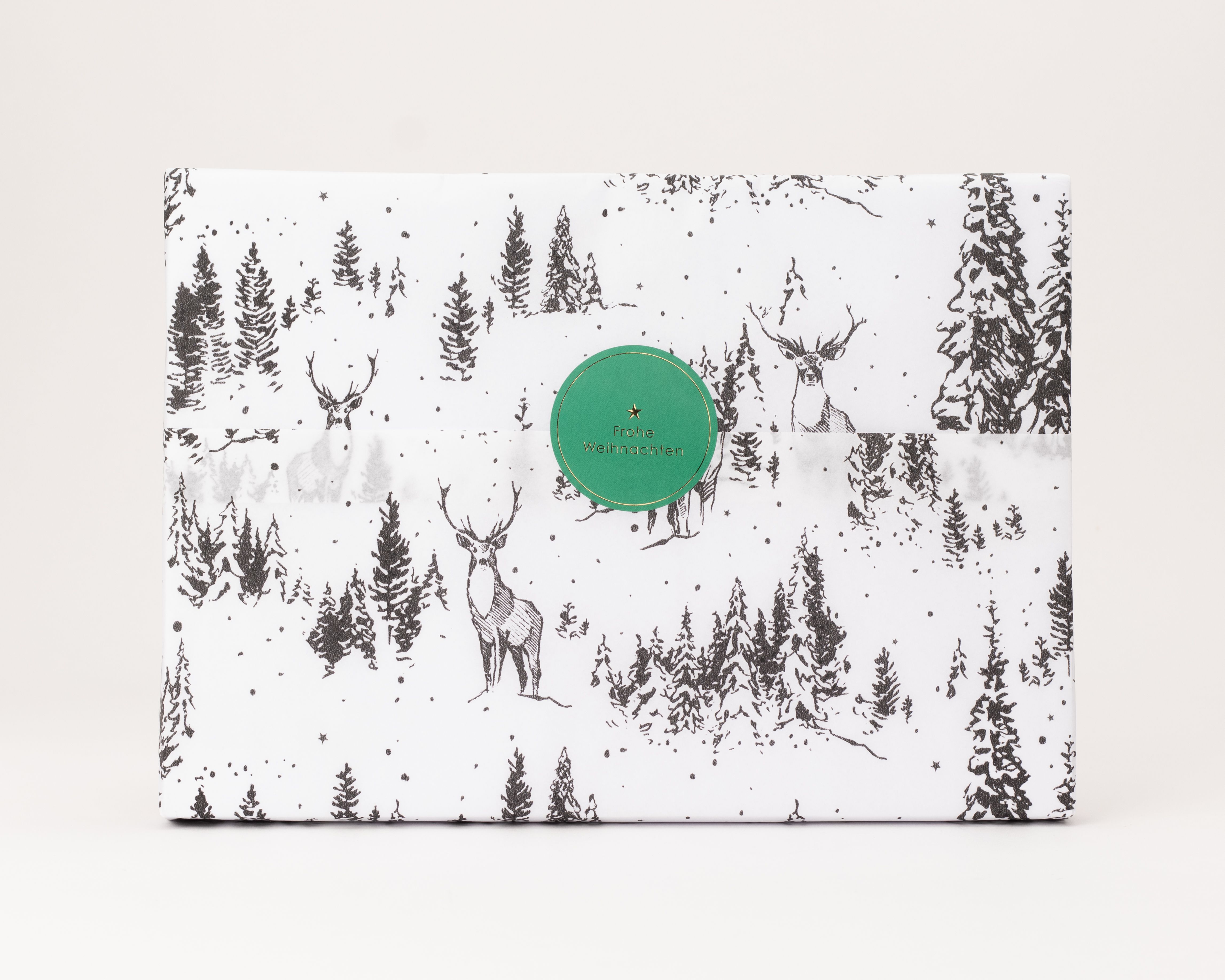 Seidenpapier weiß mit schwarzem Winterwaldmotiv und Hirschen. Darauf ein runder grüner Aufkleber mit goldener Aufschrift Frohe Weihnachten