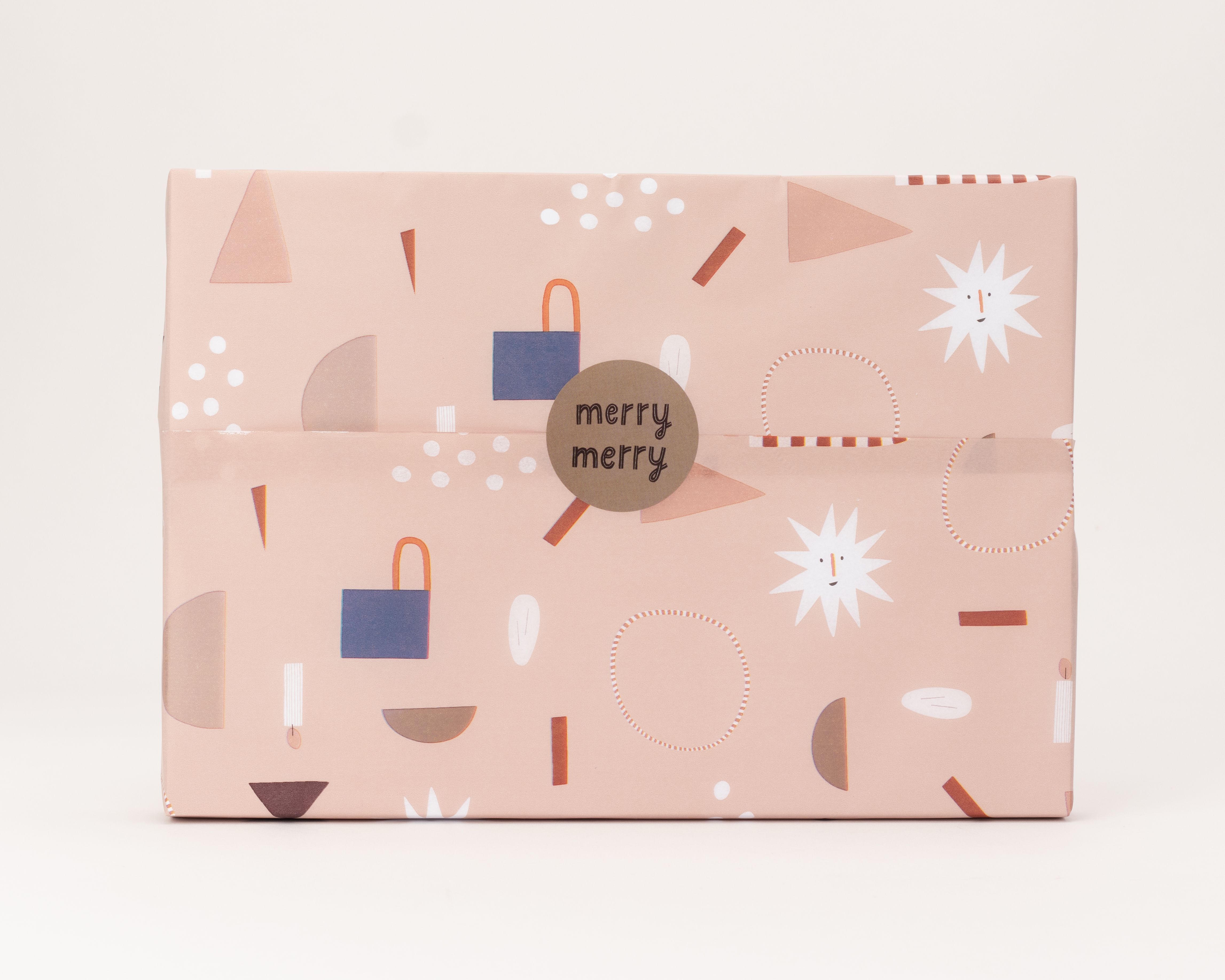 Seidenpapier rosa mit festlichem Muster aus weißen Punkten, blauen Formen und stilisierten Sternen. Darauf ein runder brauner Aufkleber mit der Aufschrift 'merry merry'