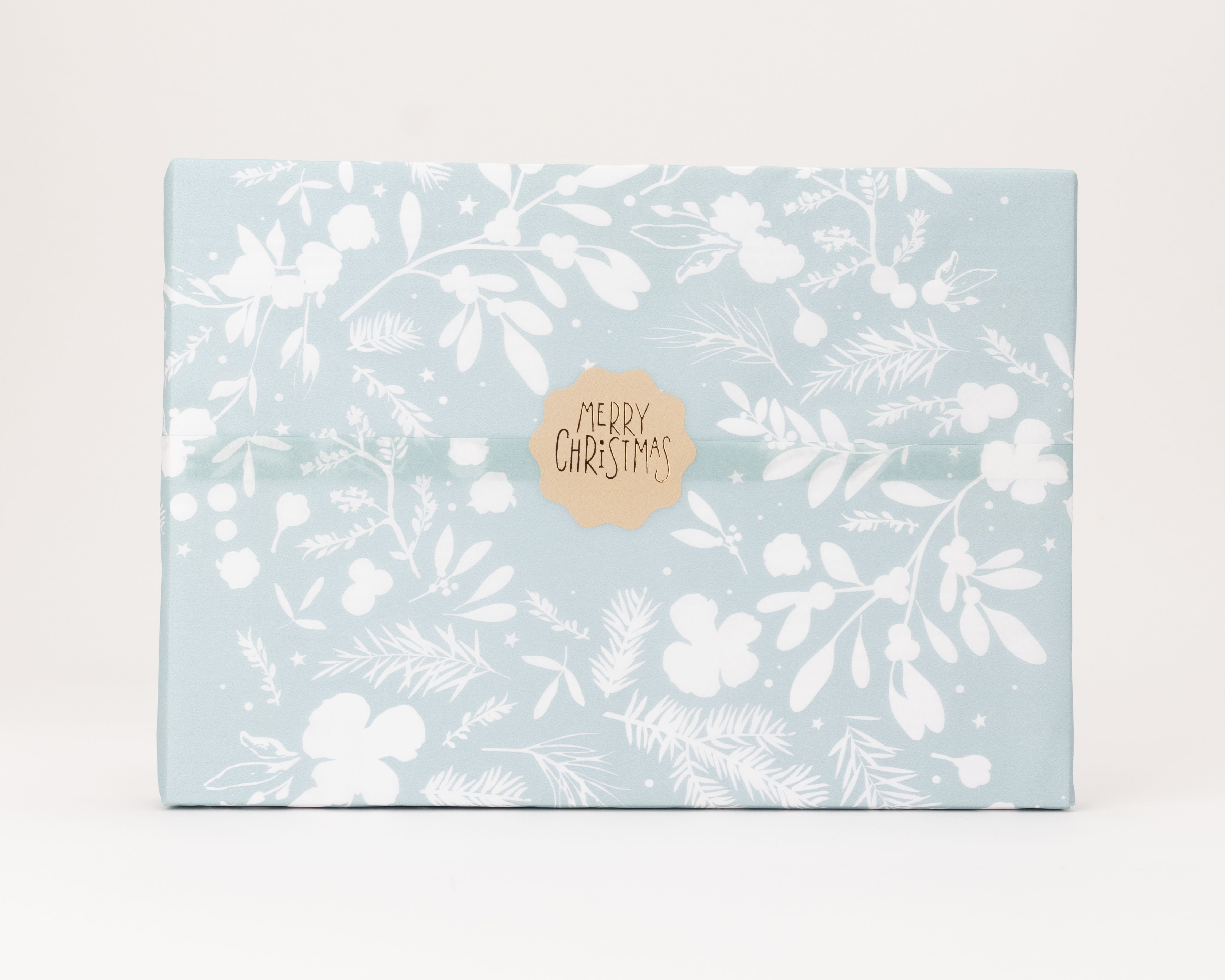Seidenpapier hellblau mit einem weißen Winter-Blumenmuster. Darauf ein brauner, gewellter Aufkleber mit der Aufschrift Merry Christmas