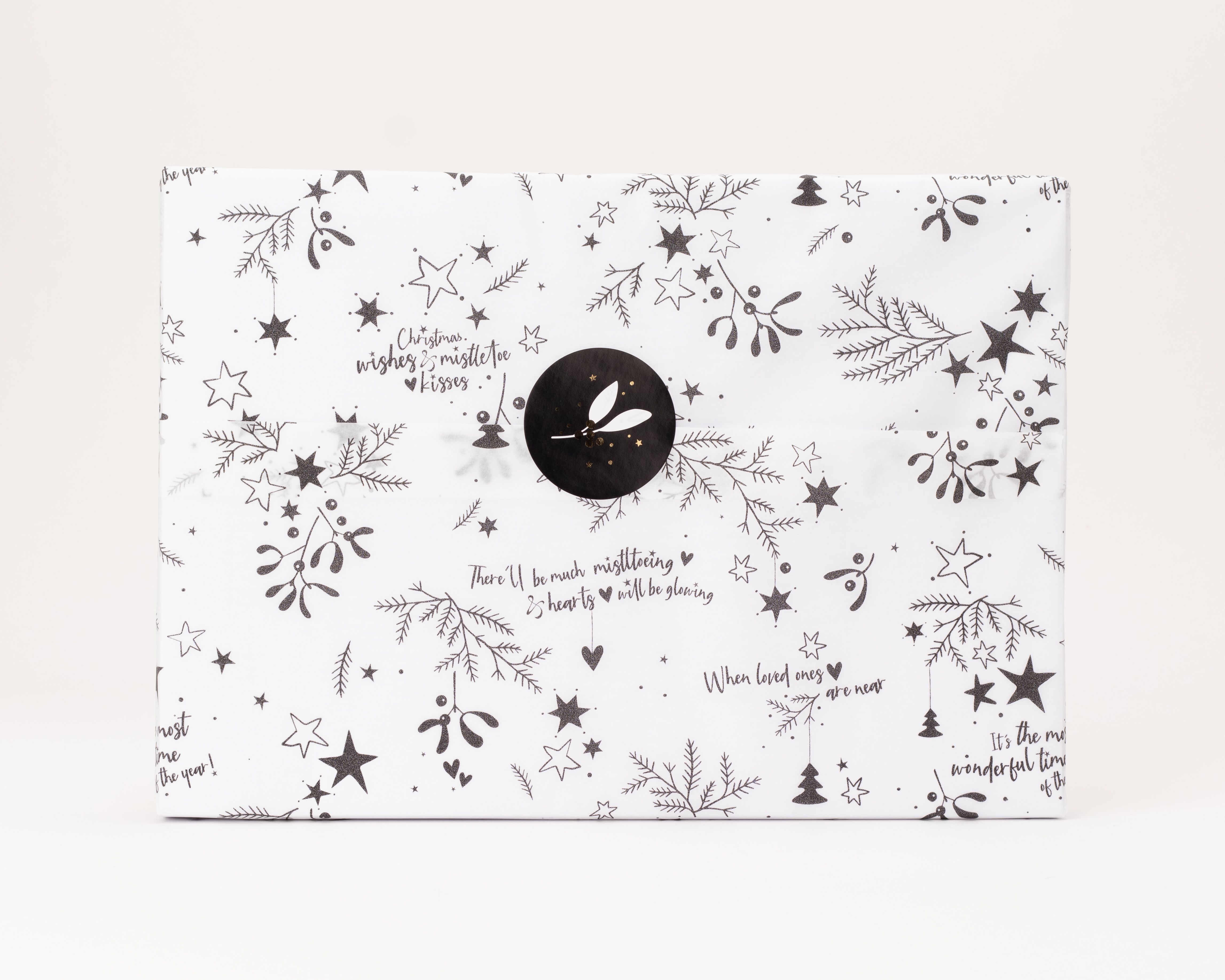 Seidenpapier weiss mit schwarzem Glitzer-Weihnachtsmotiv aus Sternen, Misteln und Schrift. Darauf ein runder schwarzer Aufkleber mit weissem Zweig und goldenen Punkten