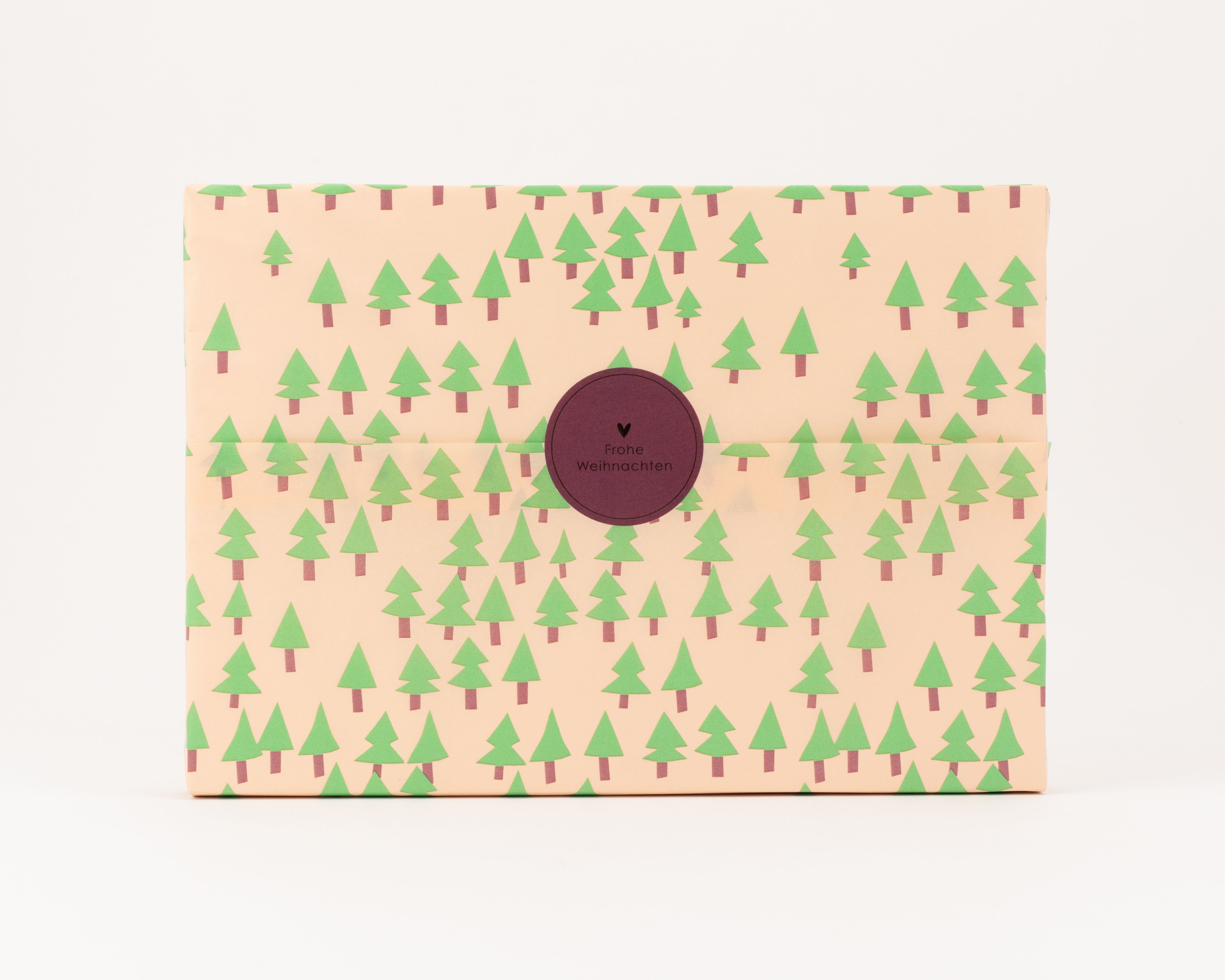 Seidenpapier mit hellbeigem Hintergrund und gruenen Tannenbaummustern. Ein dunkelroter runder Aufkleber zeigt Frohe Weihnachten
