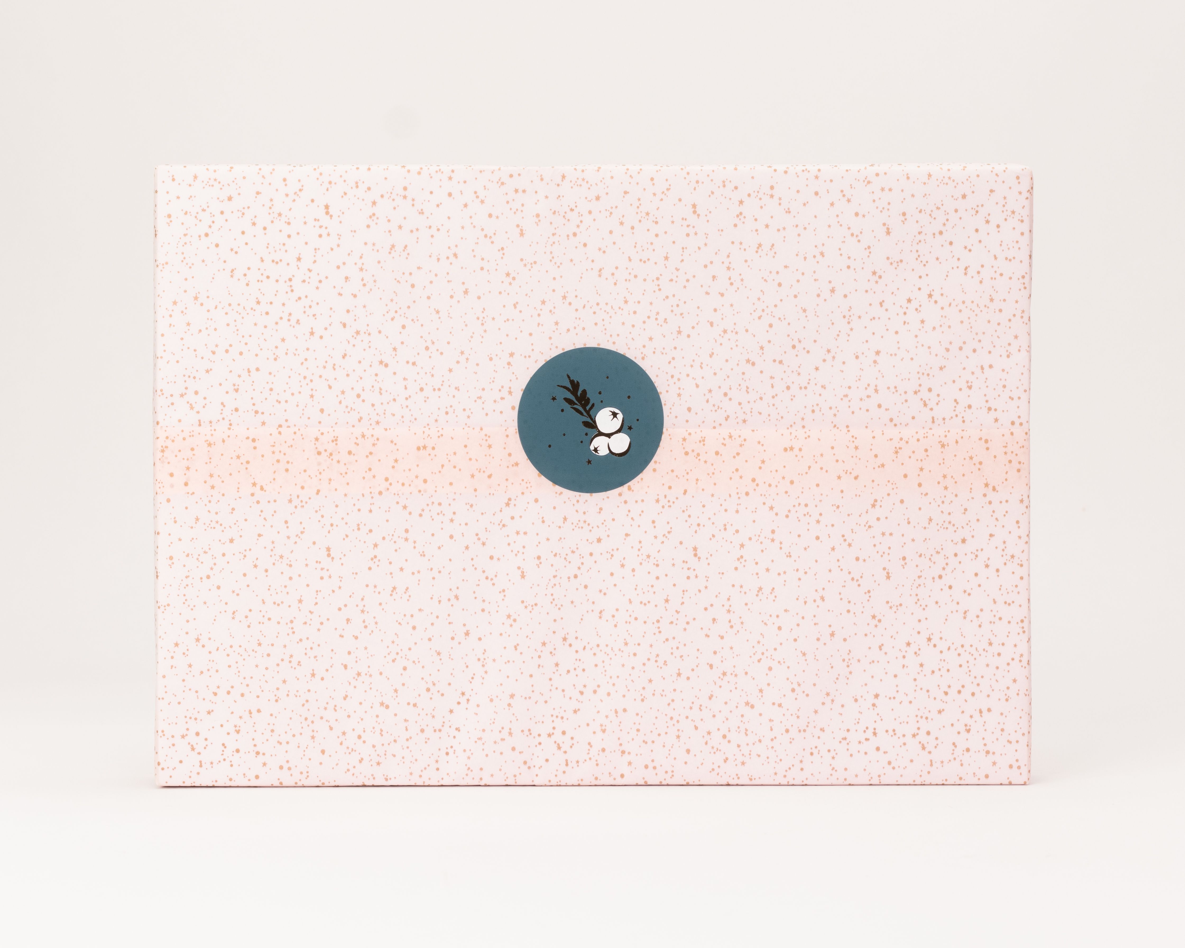 Seidenpapier in Rosa mit funkenden Sternen und Punkten in Gold. Darauf ein runder Aufkleber in Blaugrün mit weißem Zweigmotiv