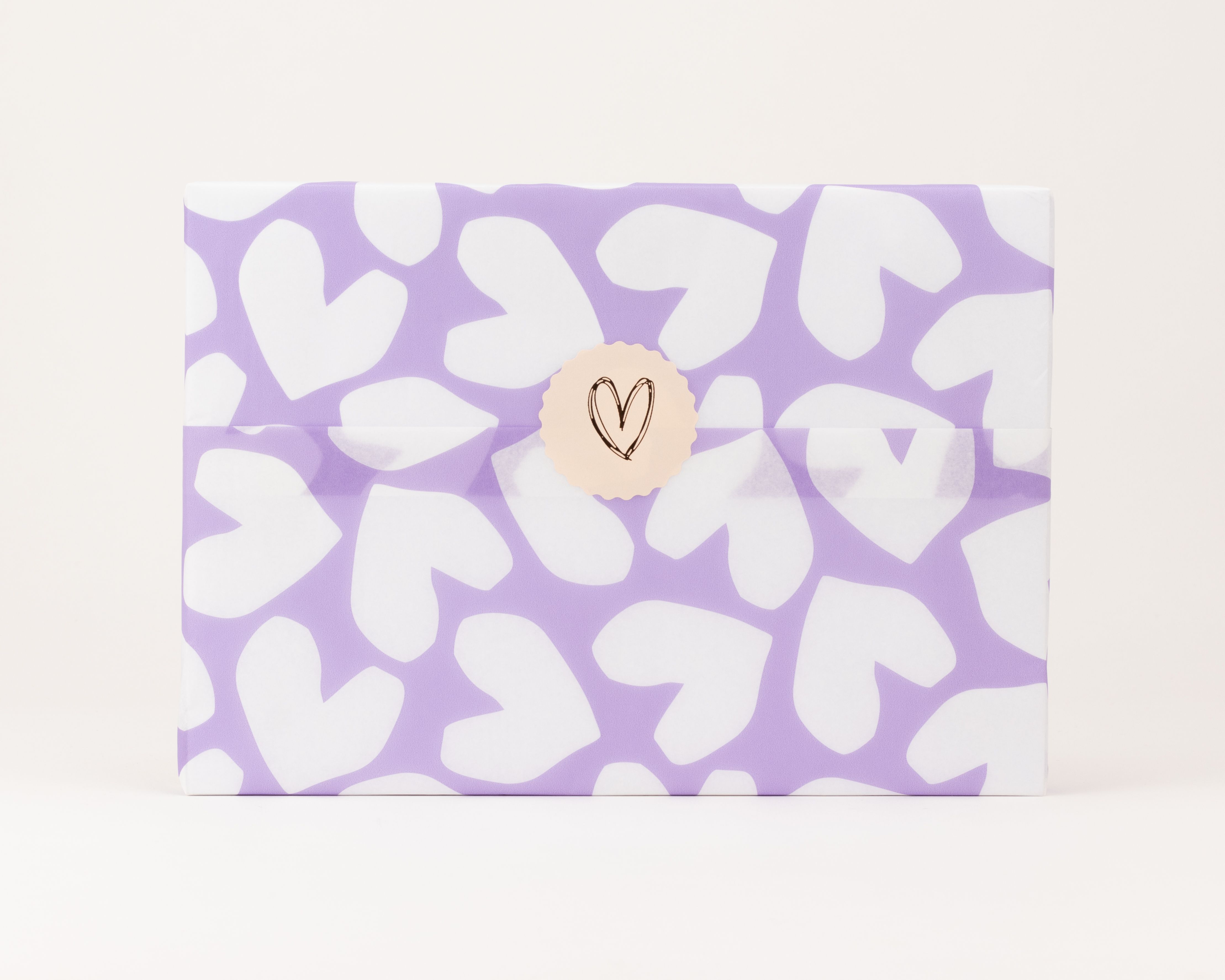 Seidenpapier mit weißen Herzen auf fliederfarbenem Grund. Darauf ein runder, beigefarbener Aufkleber mit einem schwarzen Herzmotiv