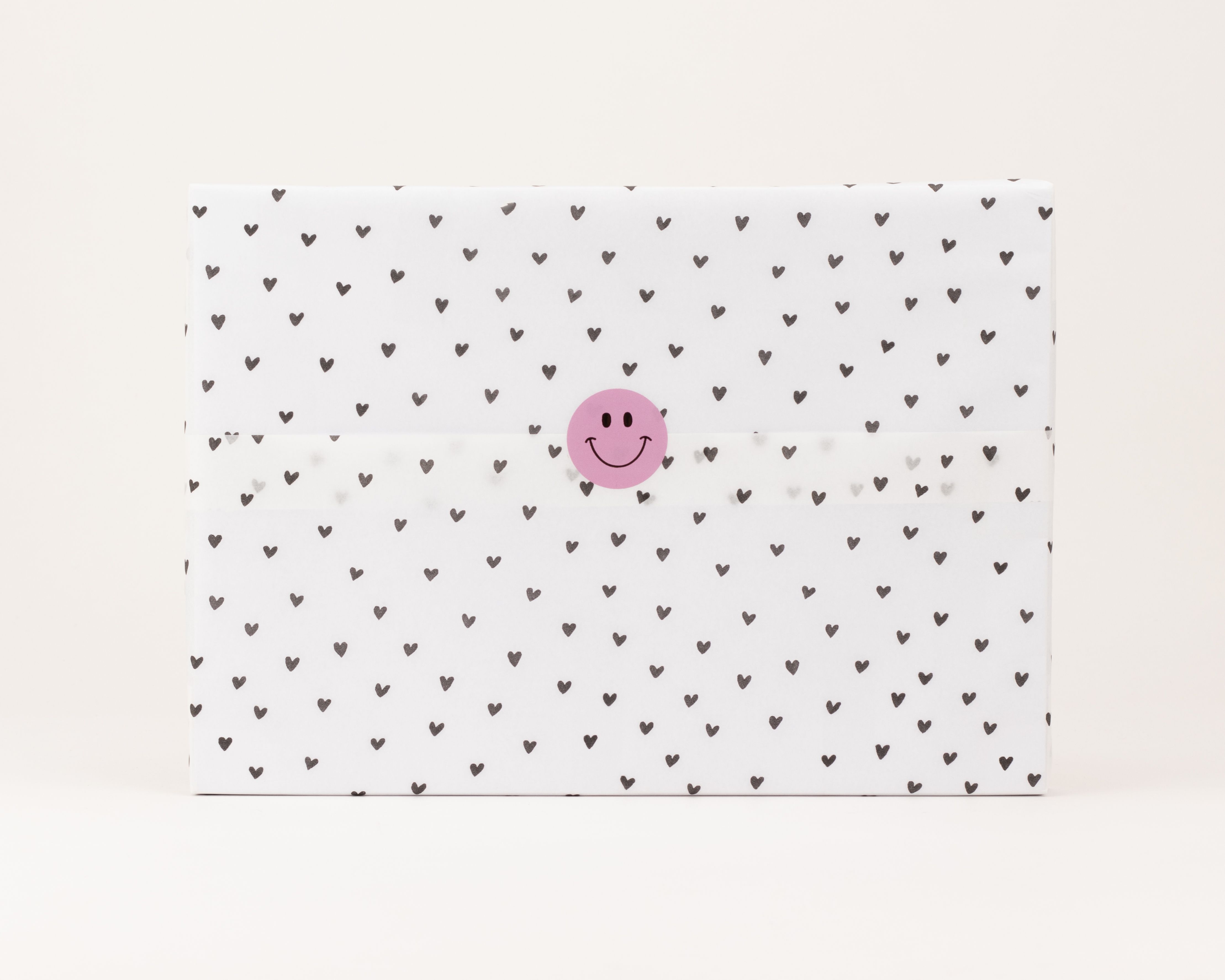 Seidenpapier in Weiß mit kleinen schwarzen Herzchen als Muster. Darauf ein runder, pinker Aufkleber mit schwarzem Smiley-Gesicht