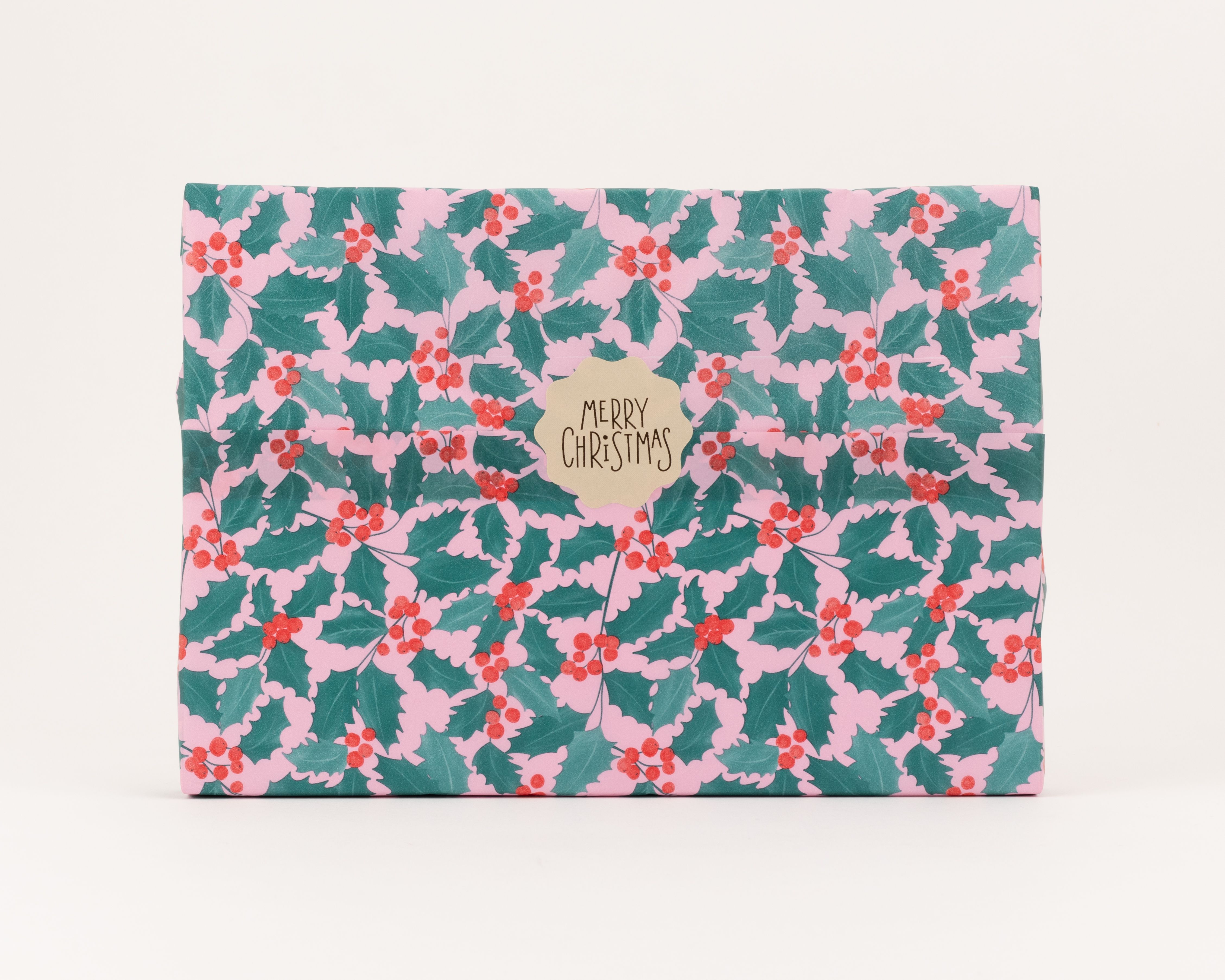 Seidenpapier mit rosa Hintergrund, grünen Ilex-Blättern und roten Beeren im Weihnachtsmuster. Darauf ein beigefarbener Aufkleber mit dem Schriftzug MERRY CHRISTMAS