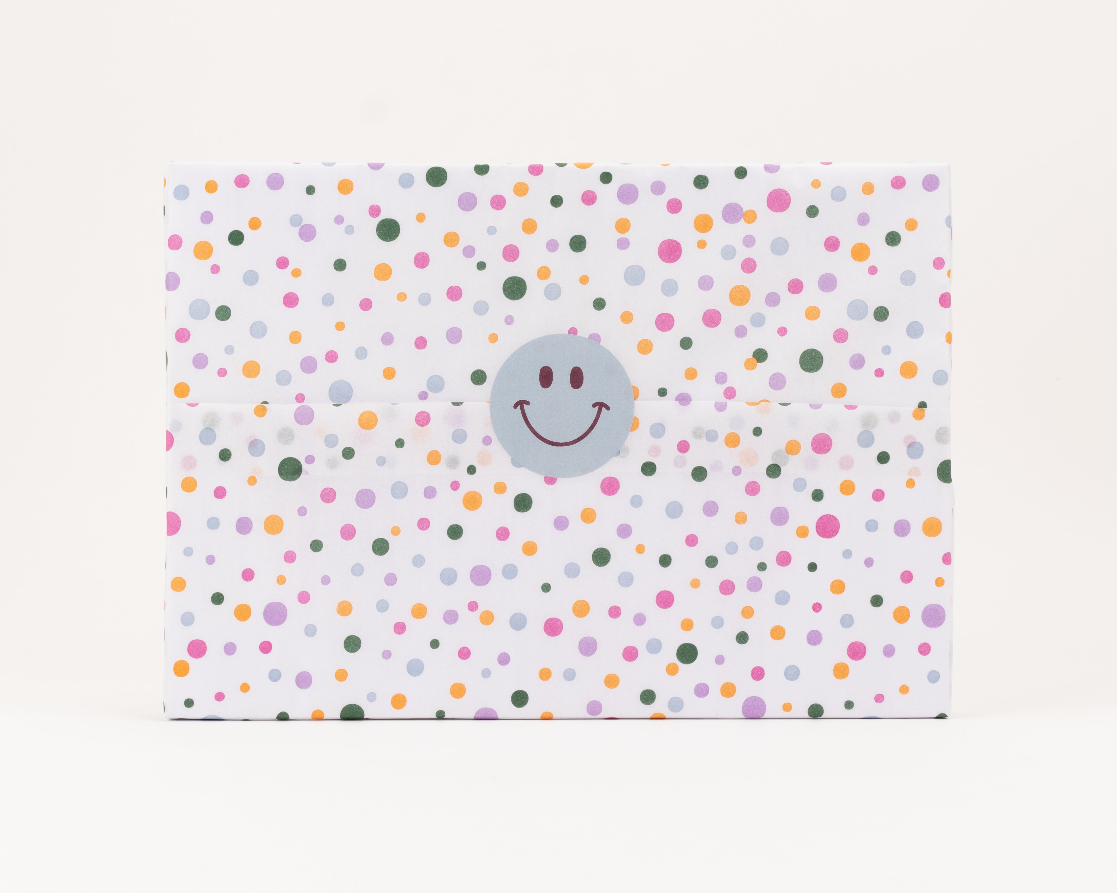 Seidenpapier in Weiß mit verschiedenfarbigen Konfettipunkten. Darauf ein runder hellgrauer Aufkleber mit einem braunen Smiley-Gesicht