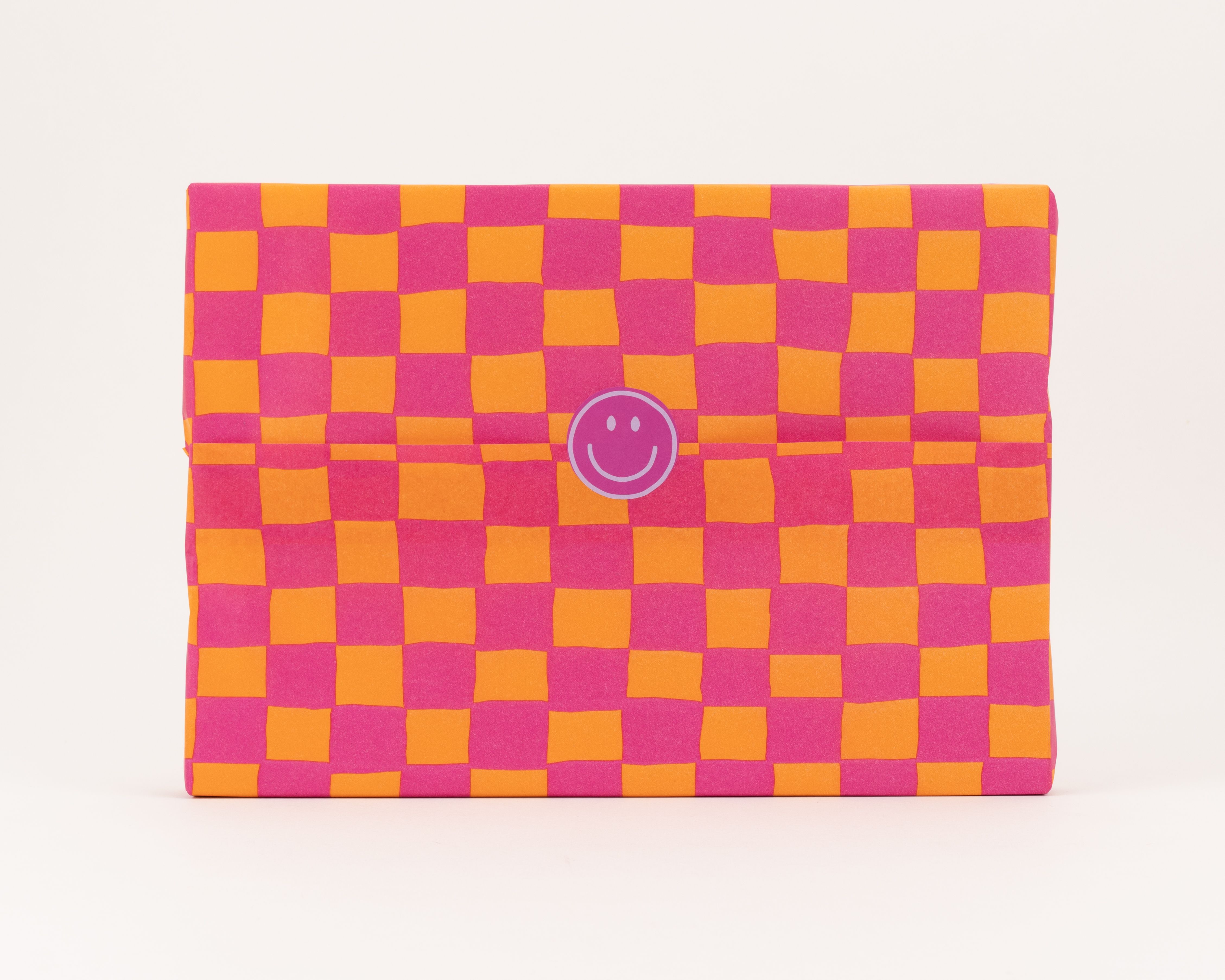 Seidenpapier mit Schachbrettmuster in kräftigem Pink und Orange. Darauf ein runder lila Aufkleber mit einem weissen Smiley Gesicht