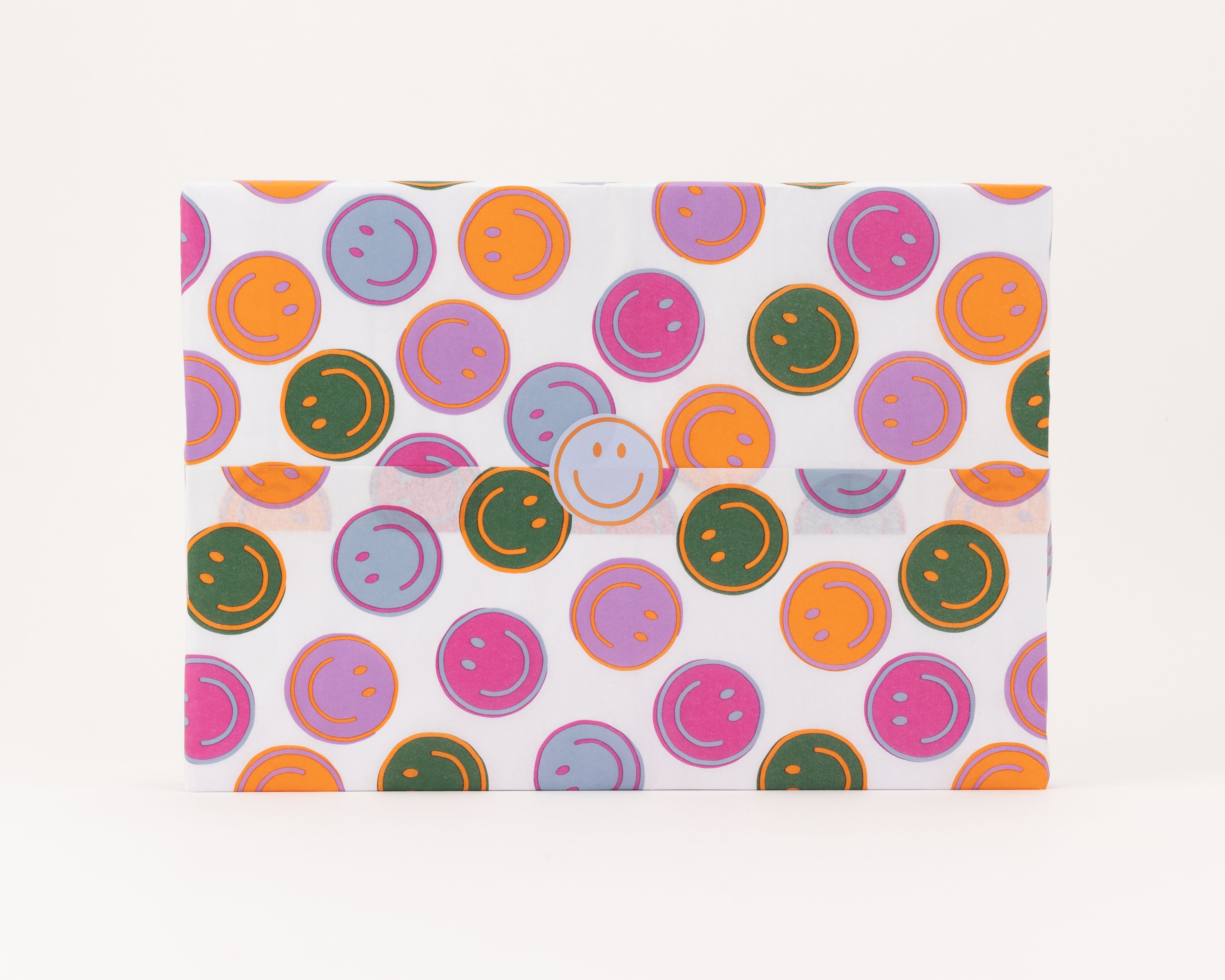 Seidenpapier in Weiß mit bunten Smileys in Grün, Pink, Hellblau, Lila und Orange, darauf ein runder hellblauer Smiley-Aufkleber