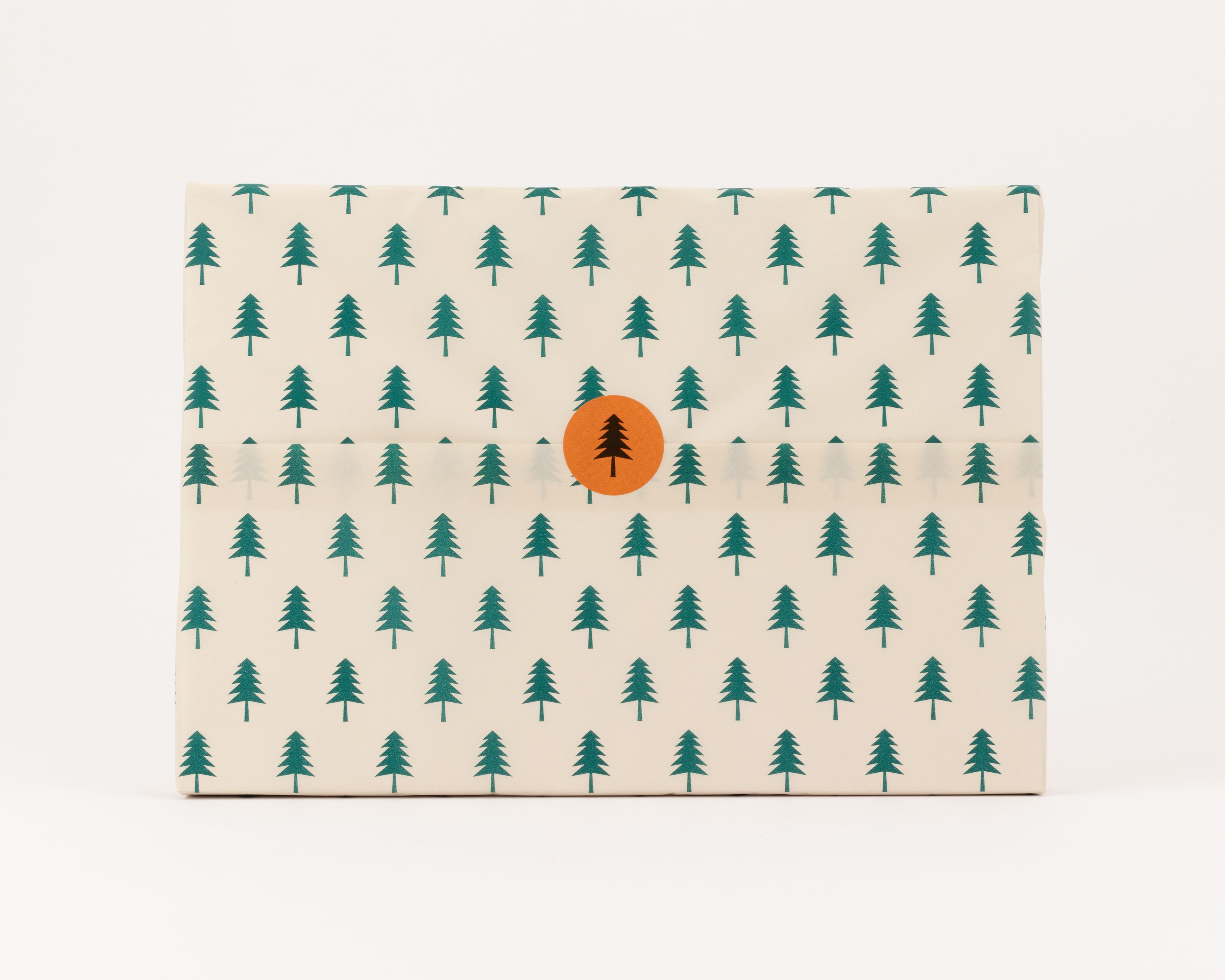 Seidenpapier mit beigem Hintergrund und einem Muster aus dunkelgrünen Tannenbäumen. Darauf ein runder, oranger Aufkleber mit einer Tannenbaum-Silhouette