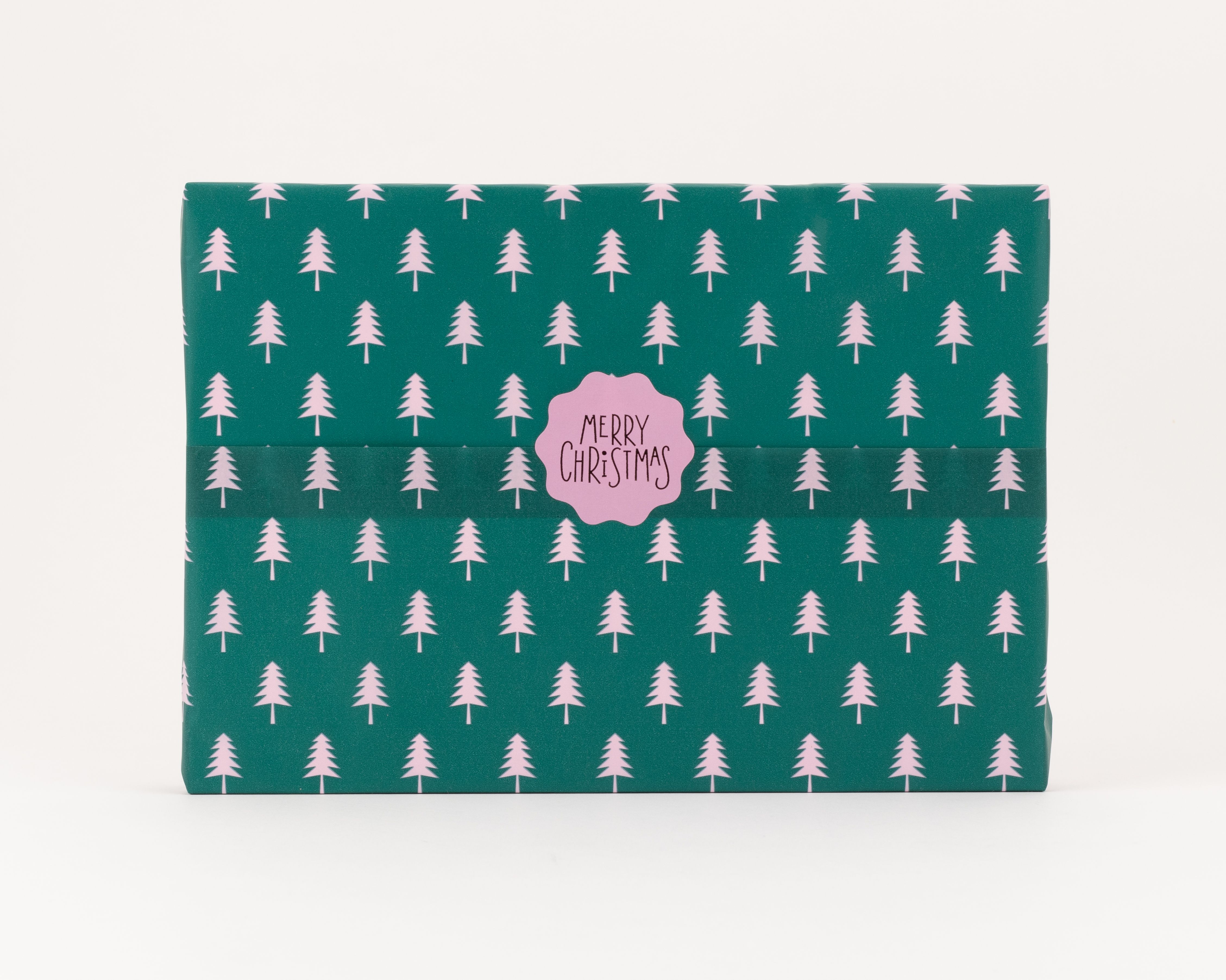 Seidenpapier in Grün mit einem hellrosa Tannenbaum-Muster. Darauf ein rosa, wellenförmiger Aufkleber mit dem Schriftzug 'MERRY CHRISTMAS'