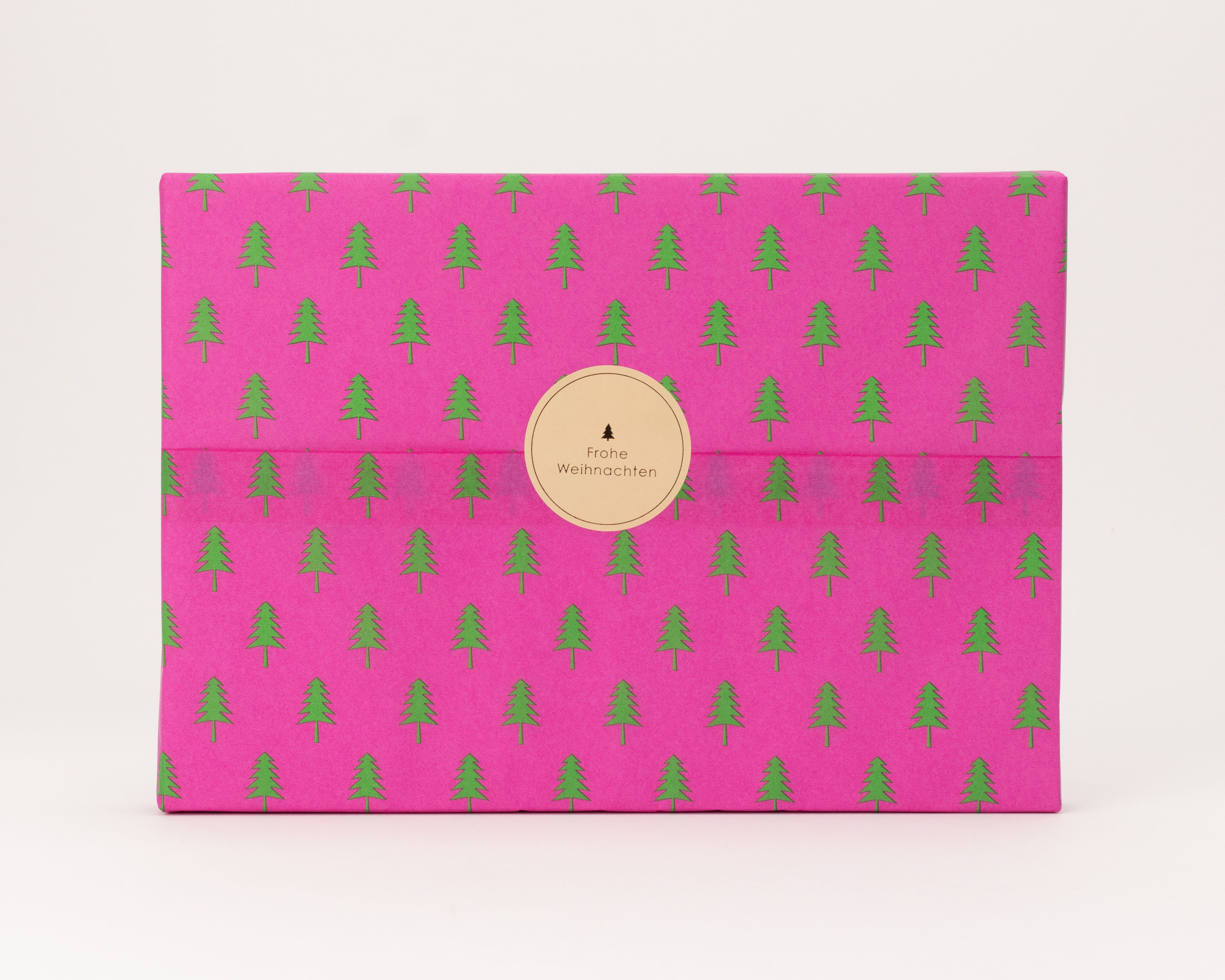 Seidenpapier pink mit grünen Tannenbäumen als Muster. Darauf ein runder beiger Aufkleber mit dem Text Frohe Weihnachten